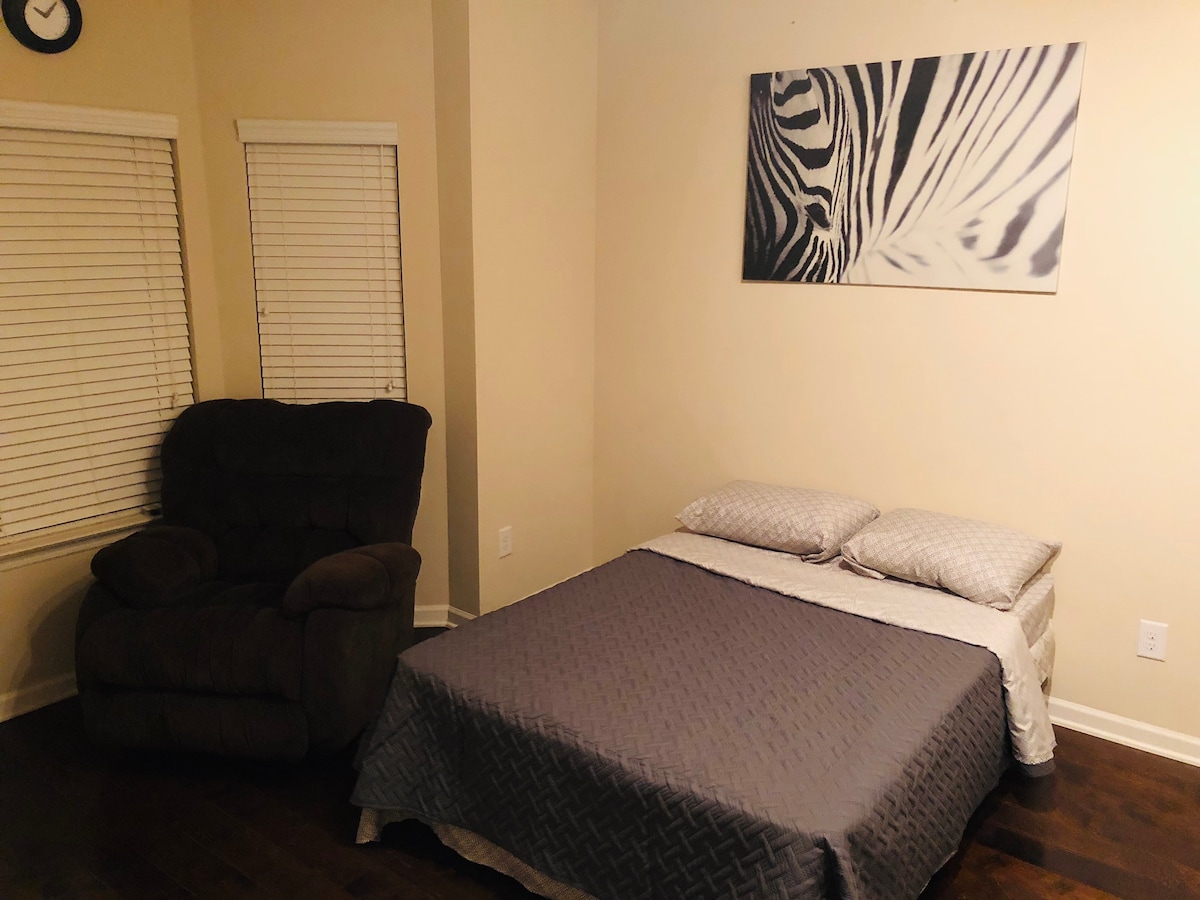 A cozy sleeping area features a spacious bed covered with a lightweight quilt and two pillows, positioned near a comfortable armchair. Natural light enters through two windows adorned with blinds, and a simple wall art piece adds a touch of decor.