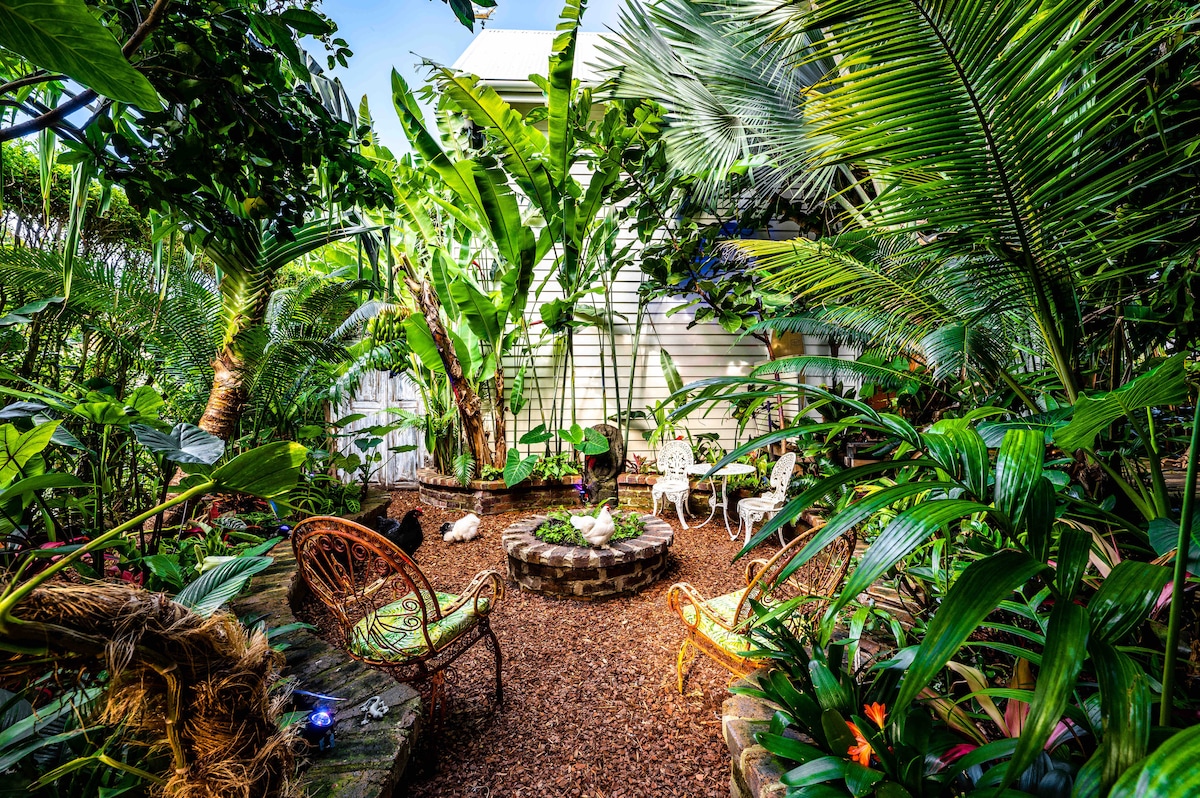 A lush private garden is surrounded by a variety of tropical plants, featuring a small seating area with wicker chairs. A circular stone fire pit is nestled among vibrant greenery, creating a serene environment. The gentle sunlight filters through the leaves, enhancing the natural ambiance.
