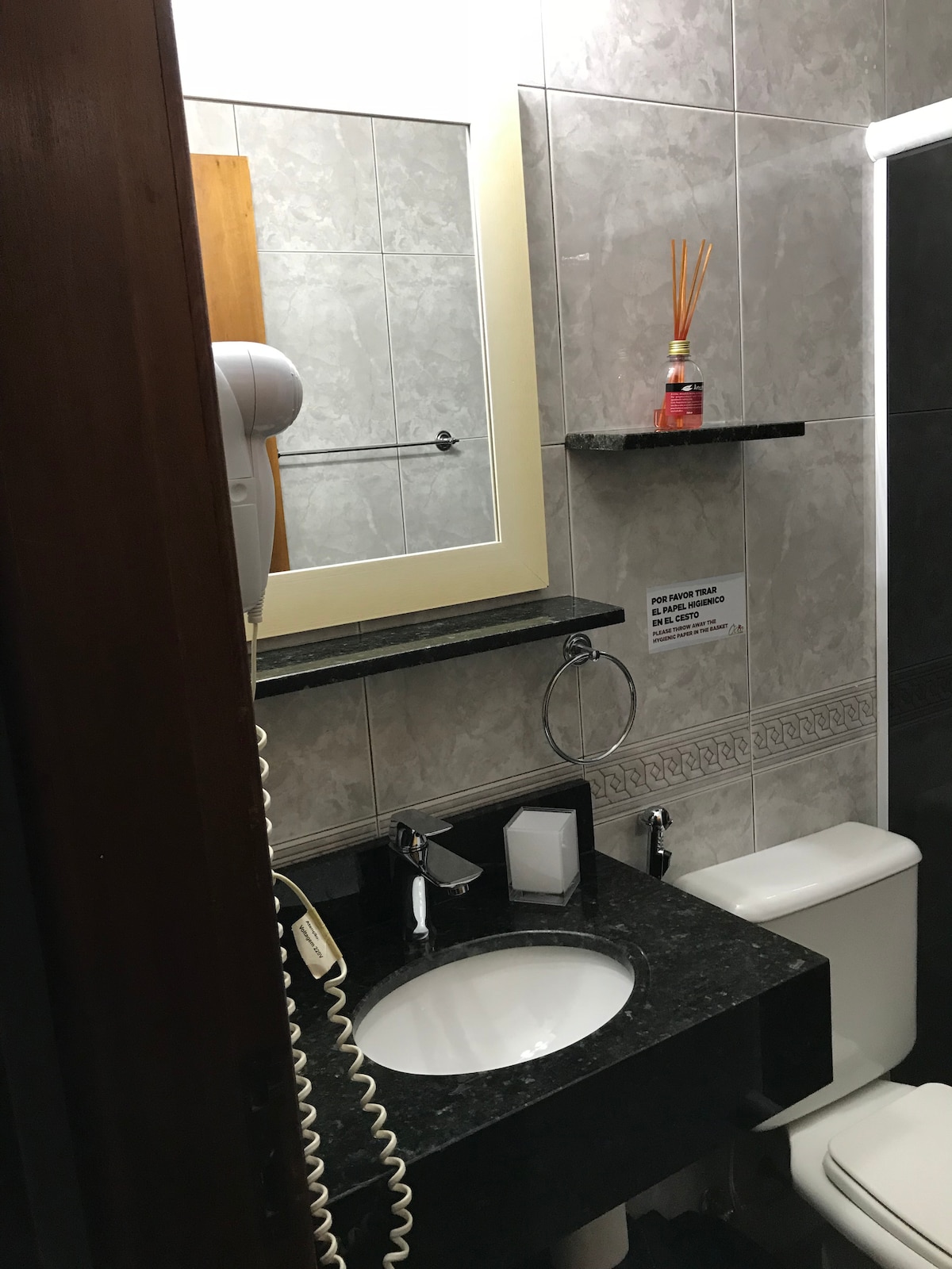 A compact bathroom features a black granite countertop with a circular sink. A large mirror is framed with a light-colored border, reflecting the soft lighting. Amenities include a small shelf with decorative reed diffusers and personal care items. The toilet is positioned adjacent to the sink.