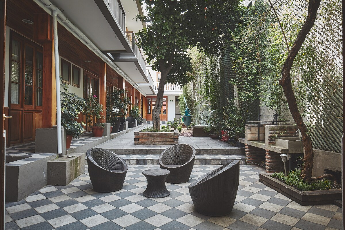A courtyard features a tiled floor with a checkerboard pattern, framed by lush greenery and planters. A set of three wicker chairs and a small table offer a welcoming seating area, surrounded by the serene atmosphere of potted plants and trees.