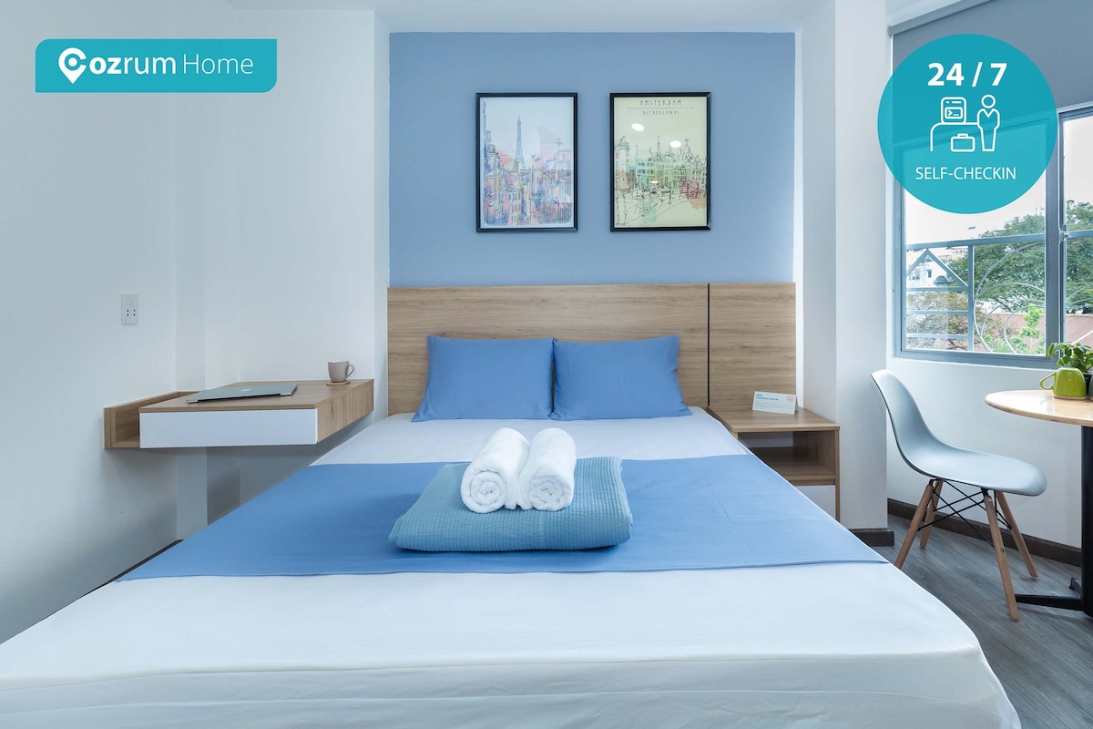 A well-lit sleeping area features a spacious bed dressed in soft blue linen, accompanied by neatly arranged towels. Wall art in muted colors is displayed above the headboard, while a small desk and chair are positioned nearby, allowing for a functional use of space.