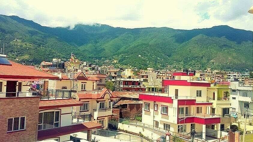 A panoramic view of a suburban neighborhood showcases a variety of well-constructed houses with red roofs. Lush green mountains rise in the background under a partly cloudy sky, enhancing the serene ambiance of the area.