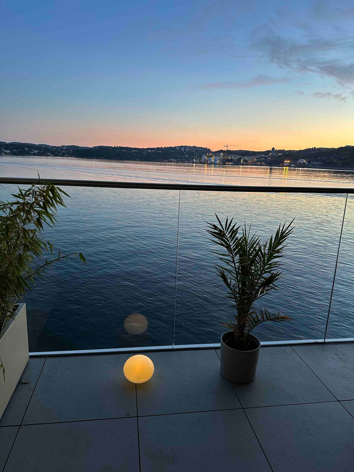 A tranquil balcony view showcases a serene body of water reflecting soft evening hues. A palm plant is positioned in a planter beside a round, glowing light, enhancing the ambiance. The glass railing offers an unobstructed view of the sunset beyond.