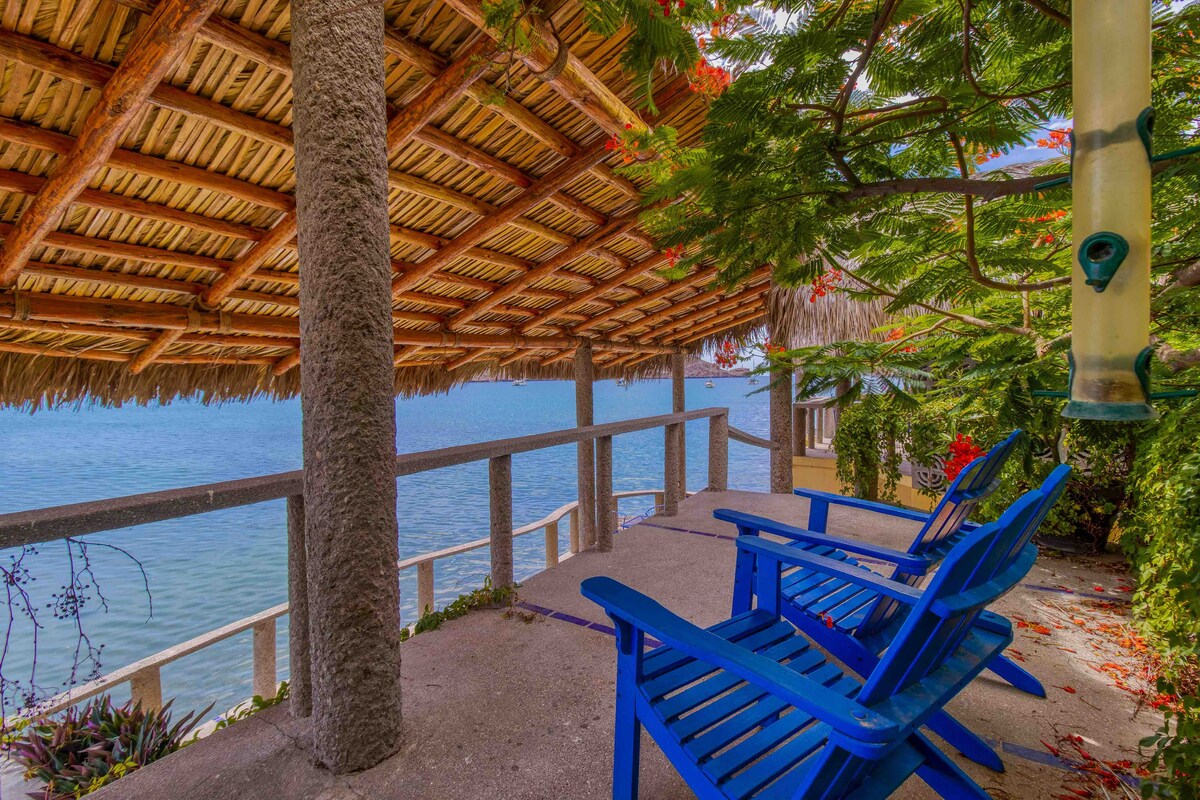 A covered patio space features two blue chairs placed on a concrete floor. The ceiling is adorned with a thatched roof, providing shade. A view of the water is visible beyond the patio, complemented by surrounding greenery and flowering plants.