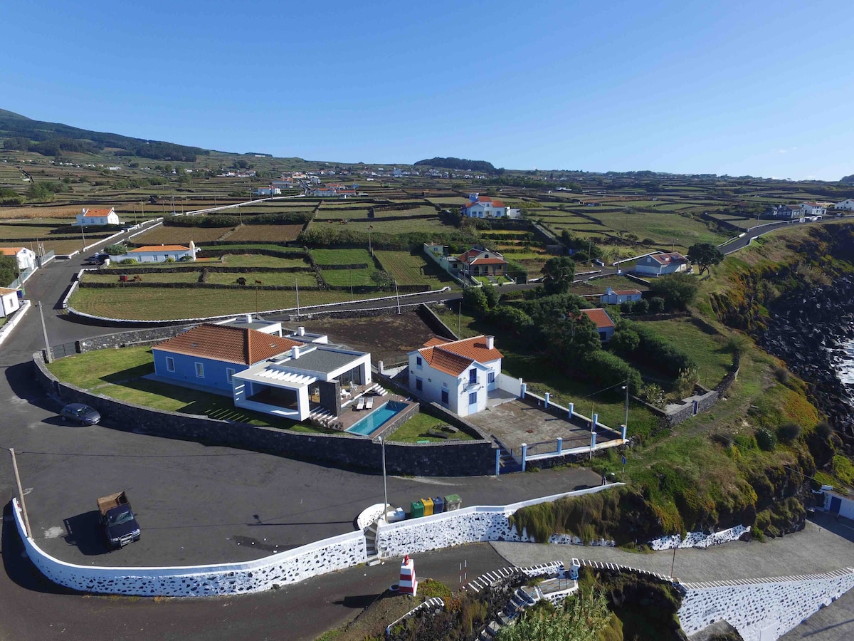 Aerial view of a coastal property nestled in lush green fields. The house displays a white exterior with a red roof, accompanied by an open patio area and a pool. Surrounding roads and countryside provide a serene backdrop with distant hills visible in the background.
