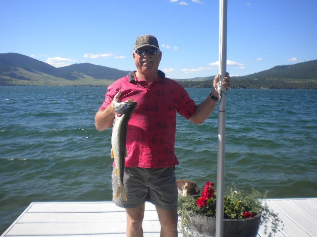 A person stands on a deck by Flathead Lake, holding a sizable fish. The serene water and surrounding hills are visible in the background under a clear blue sky. Red flowers are seen in a planter nearby, adding a touch of color to the scene.
