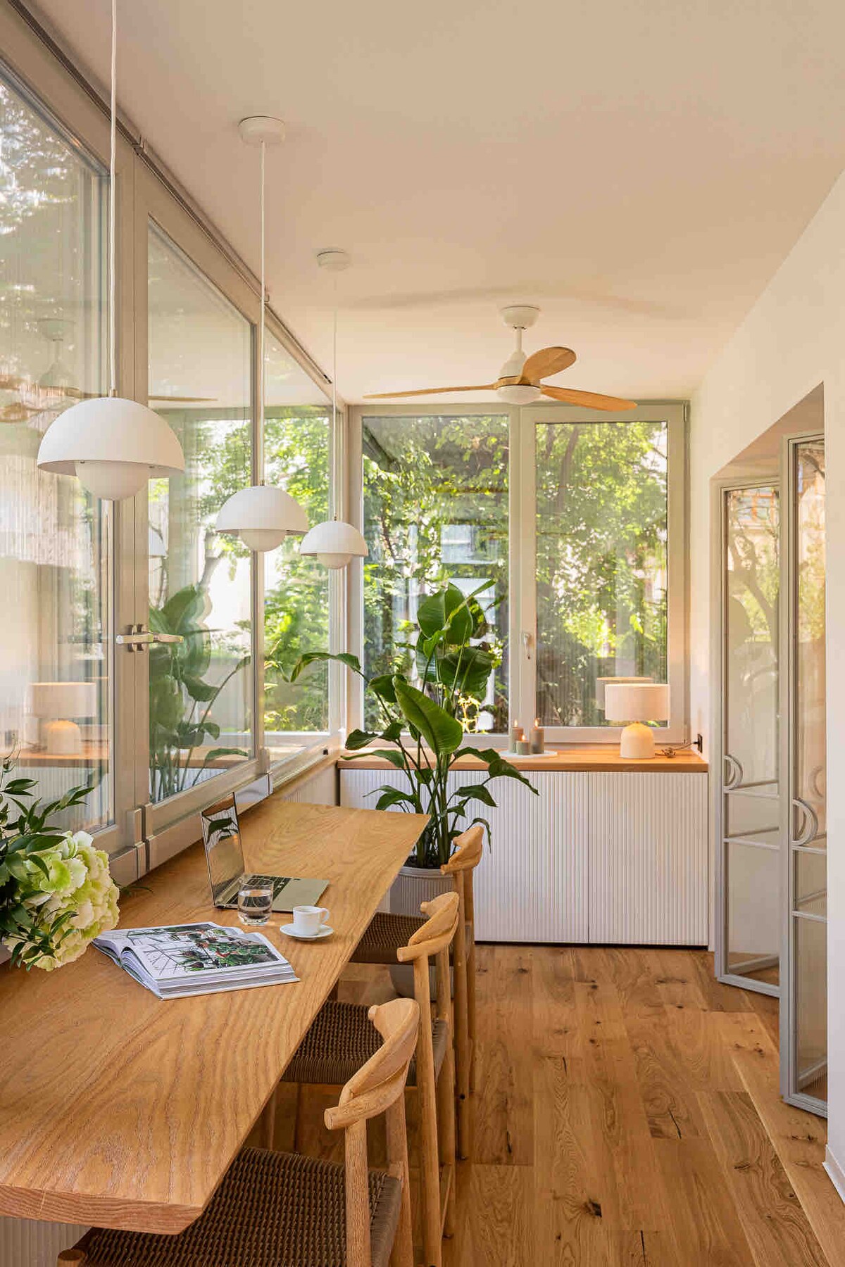 A bright and airy space features a wooden table with chairs, complemented by large windows that bring in natural light. Potted plants add greenery, while pendant lights hang above, creating a serene atmosphere. A small decorative lamp is placed on a nearby surface.