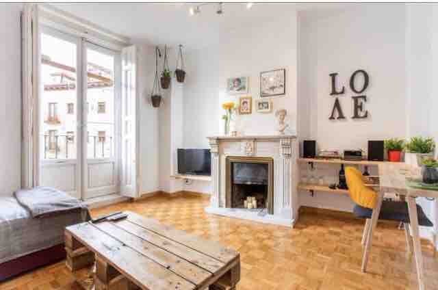 A bright living area features large windows with views of the street, complemented by a comfortable seating area and a wooden coffee table. A decorative fireplace is positioned centrally, with a small music setup and plants adorning the mantel and shelves.