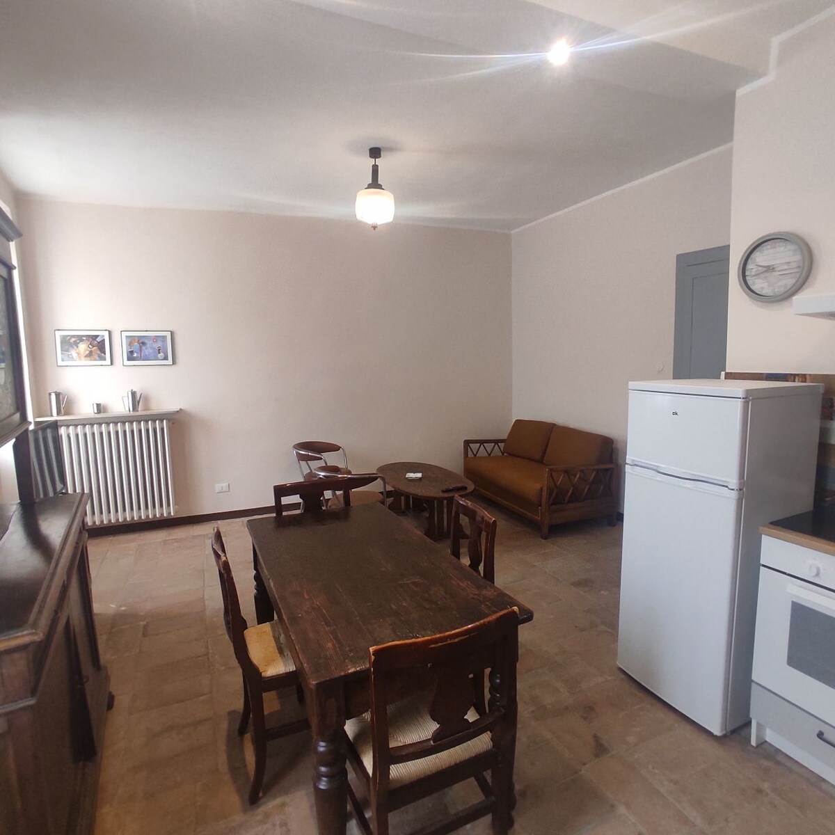 An open space is depicted, featuring a wooden dining table surrounded by four chairs. A brown sofa is positioned nearby, and a white refrigerator and oven are visible in the kitchen area. Natural light illuminates the room, enhancing the neutral tones of the walls.