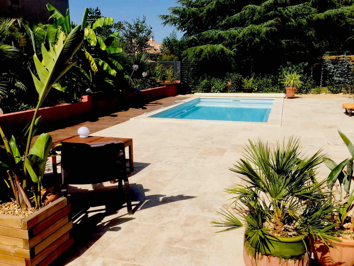 The outdoor space features a private pool surrounded by a stone patio. Lush greenery, including banana plants and palm trees, provides a tropical feel. A wooden table and chairs are positioned nearby, offering a relaxing spot to enjoy the setting.