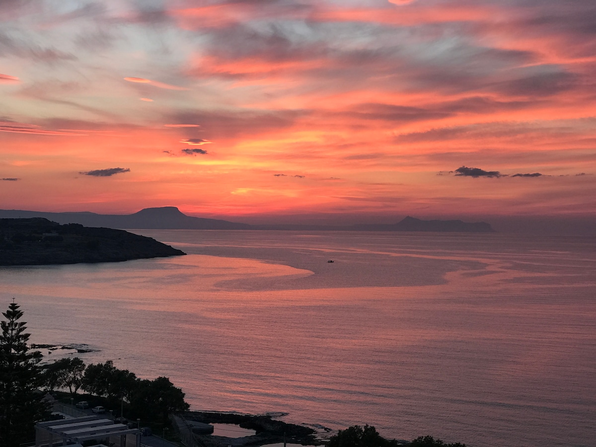 A serene sunset paints the sky in shades of pink and orange over the calm Cretan Sea. Silhouetted hills can be seen in the distance, while the water reflects the vibrant colors. The scene captures the tranquil beauty of the surrounding landscape.