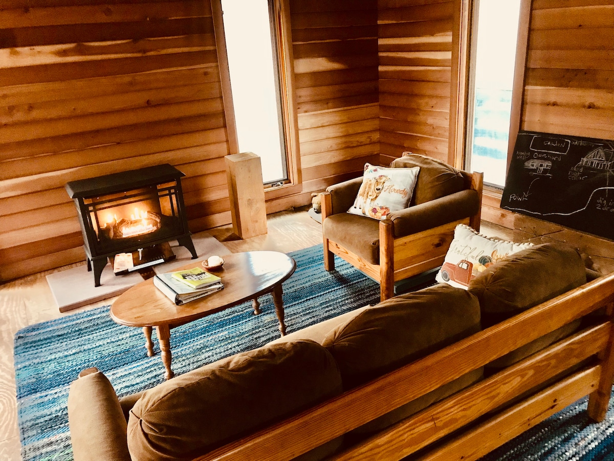 The interior features a comfortable seating area with a plush couch and wooden chairs arranged around a small coffee table. A wood-burning stove is positioned against a wooden wall, providing warmth. Natural light filters through nearby windows, illuminating the cozy atmosphere.