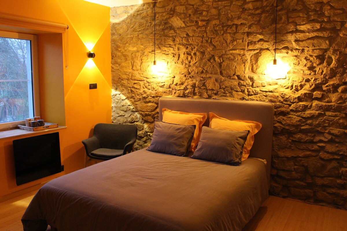 A cozy bedroom features a spacious bed with soft gray bedding and orange accent pillows. Stone walls are complemented by modern lighting fixtures on either side. A comfortable chair is positioned near a window, allowing natural light to accentuate the warm tones of the room.