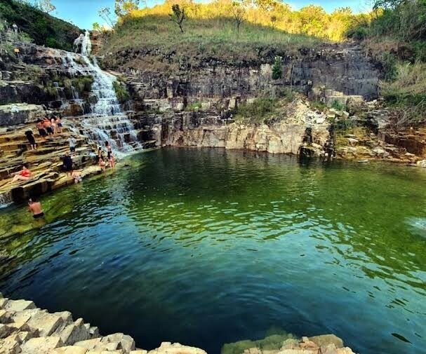 A natural pool is surrounded by rugged rock formations, with gentle waterfalls cascading into the green water below. Visitors can be seen enjoying the serene environment, and vibrant greenery peeks through the rocky terrain, enhancing the tranquil setting.