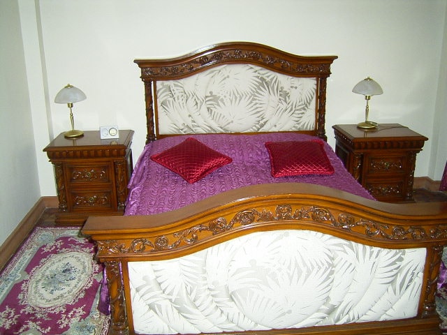 A wooden bed frame with intricate carvings is featured, showcasing a spacious bed adorned with a purple bedspread. Two matching bedside tables hold elegant lamps, and decorative red pillows are placed on the bed. A patterned area rug adds warmth to the room.