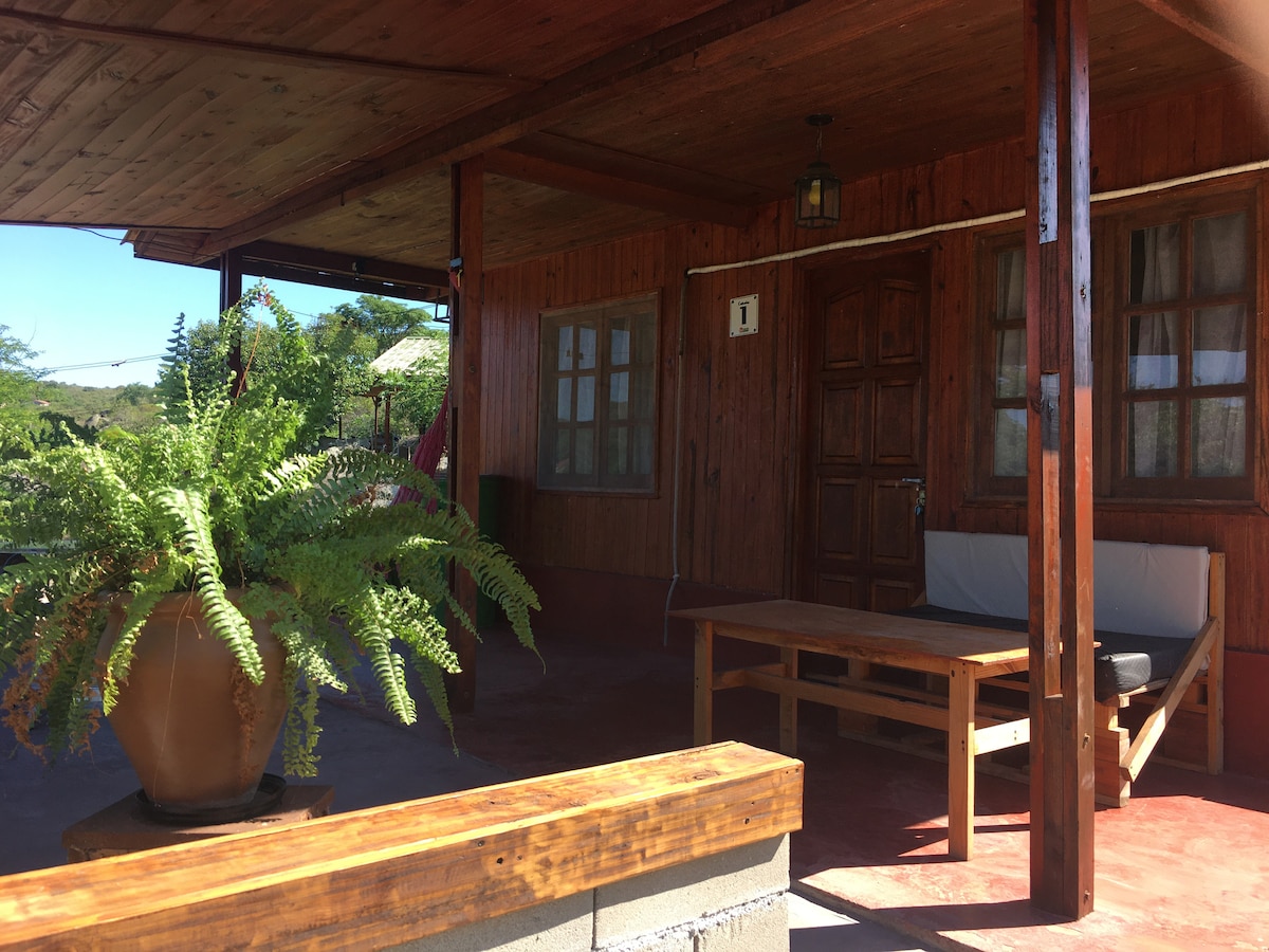 An outdoor seating area features a wooden table and benches, accompanied by a large potted fern. The front of the wooden cabin includes several windows and a main entrance door, all set against a backdrop of natural surroundings.