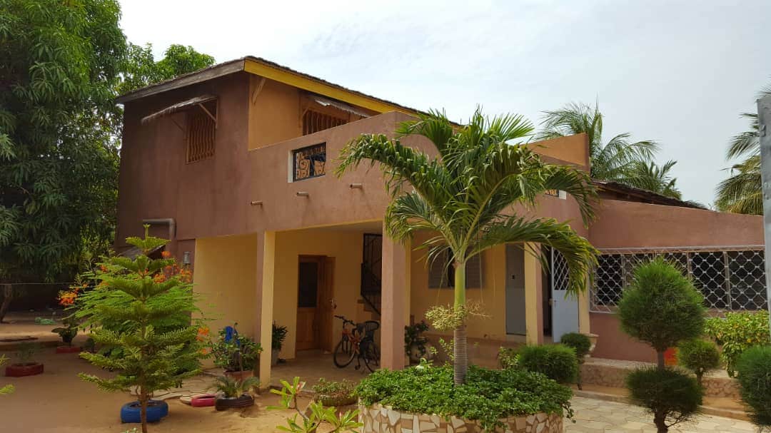The exterior of the house features a combination of warm earth tones with a yellow accent on the upper level. Lush greenery surrounds the property, including palm and other tropical plants. An inviting entrance is visible, leading to the upper floor balcony while bicycle storage is situated nearby.