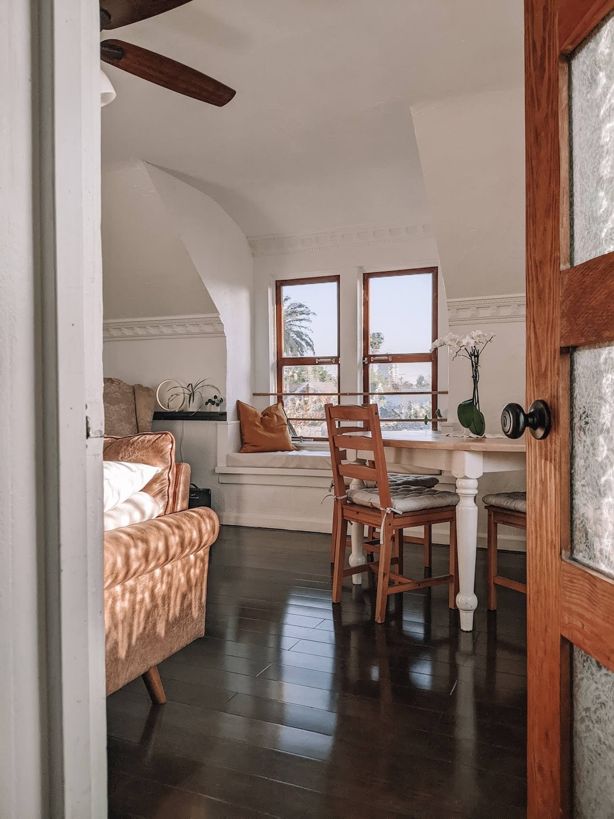 A bright and airy living space is visible through an open door. Large windows allow natural light to illuminate the hardwood floors. A table with chairs sits adjacent to a cozy seating area, featuring plants and decorative cushions, creating a welcoming atmosphere.