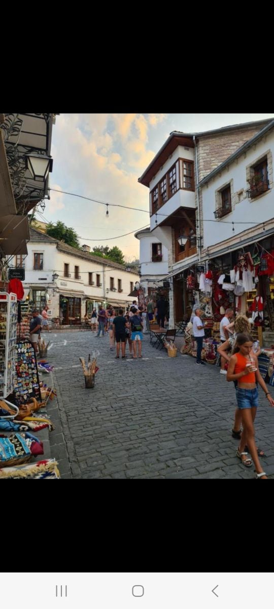 A vibrant street scene captures visitors exploring local shops in the Old Bazaar. Colorful merchandise is displayed prominently along the cobblestone path. Traditional architecture is highlighted by white buildings, with some featuring wooden balconies. The image conveys a lively atmosphere filled with people engaging with the surroundings.