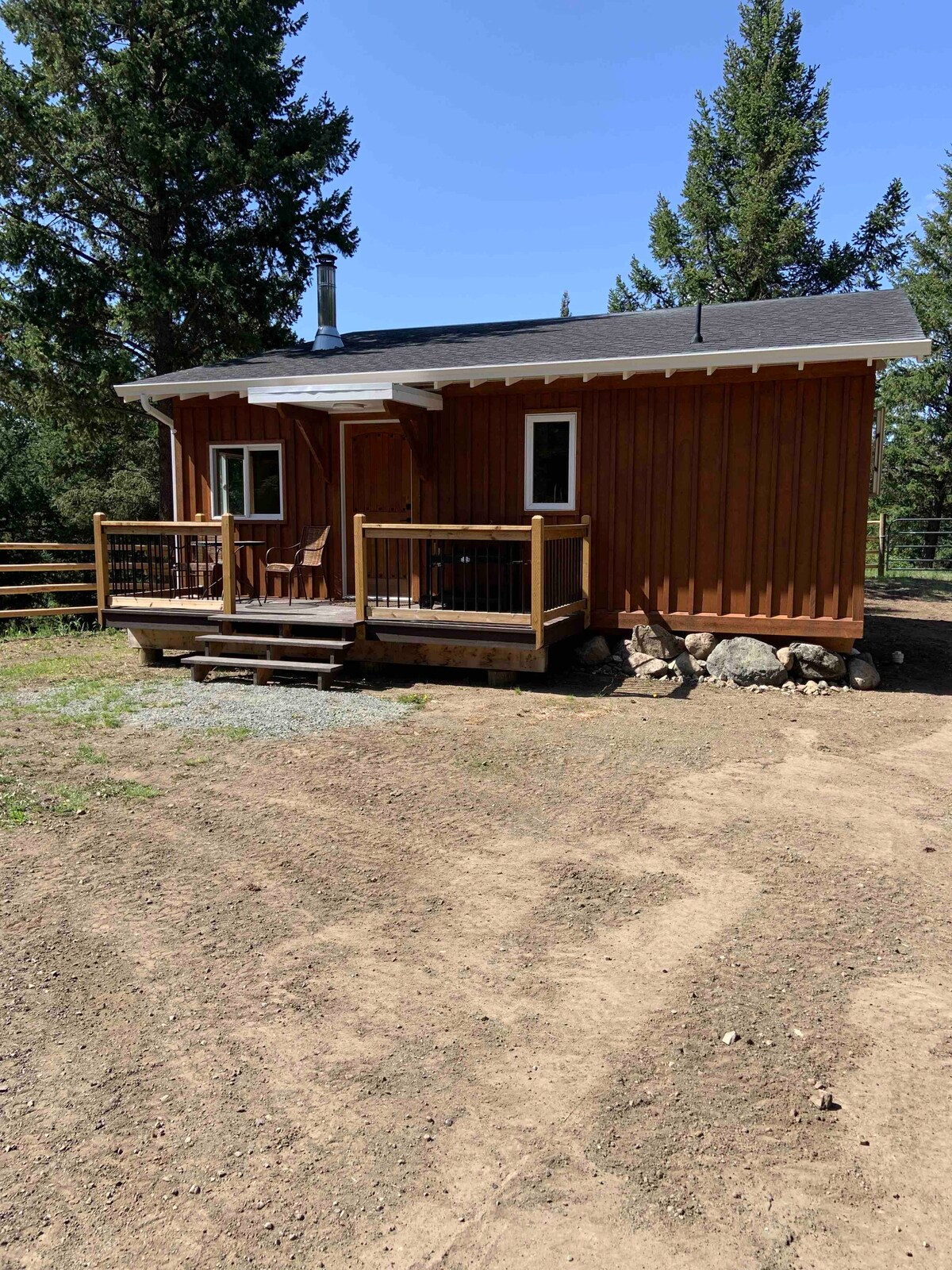 The exterior of the cabin is showcased, featuring warm wooden siding and a sloped roof. A front porch is visible, with seating provided by a pair of chairs. Surrounding trees enhance the natural setting, while a gravel pathway leads up to the entrance.
