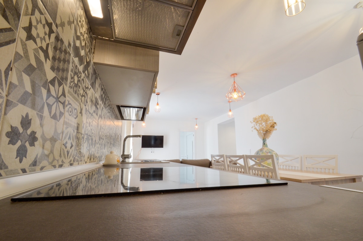 A modern kitchen showcases a sleek design with a glossy countertop and an induction cooktop. Decorative tiles in various patterns adorn the wall, adding an artistic touch. Soft lighting is provided by hanging fixtures, creating a bright and functional cooking space.