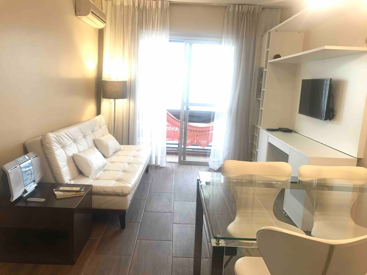 A living area is shown with a white sofa bed positioned beside a wooden side table. A glass dining table with white chairs is visible, complementing the light-colored decor. Large windows allow natural light to fill the space, with sheer curtains and a glimpse of a balcony visible in the background.