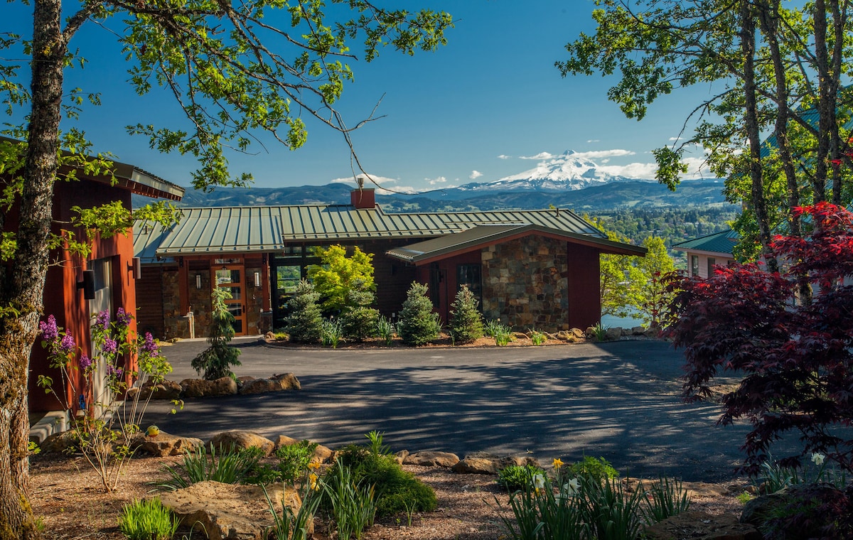 The exterior of a modern home is presented amid lush greenery, showcasing a well-maintained driveway. A mountain view is visible in the background, with trees framing the scene and complementing the contemporary architecture.