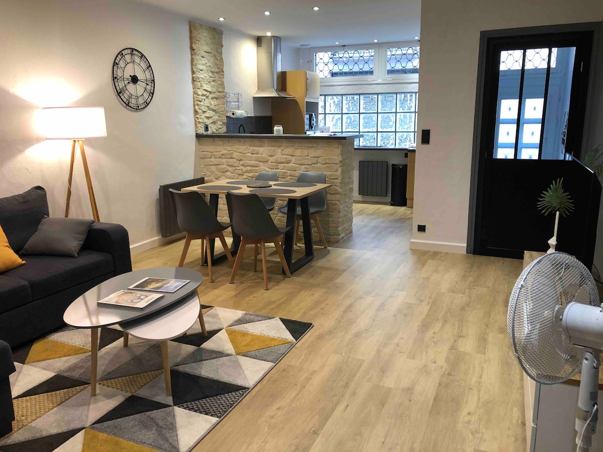 An open living space features a cozy dark sofa beside a round coffee table. A dining area with a wooden table and gray chairs sits opposite a modern kitchen with stone accents. Natural light filters in through large windows, creating a warm ambiance.