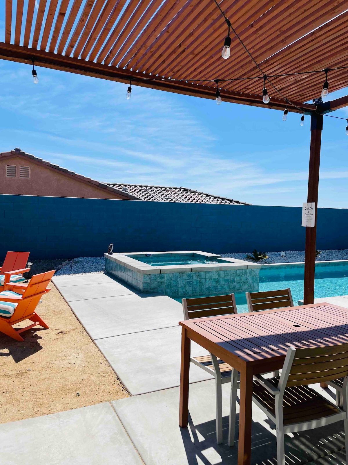 An outdoor space features a dining table with several chairs under a shaded pergola, next to a saltwater pool and spa. Colorful adirondack chairs are positioned nearby, and a blue wall provides privacy. String lights are visible above, adding gentle illumination.
