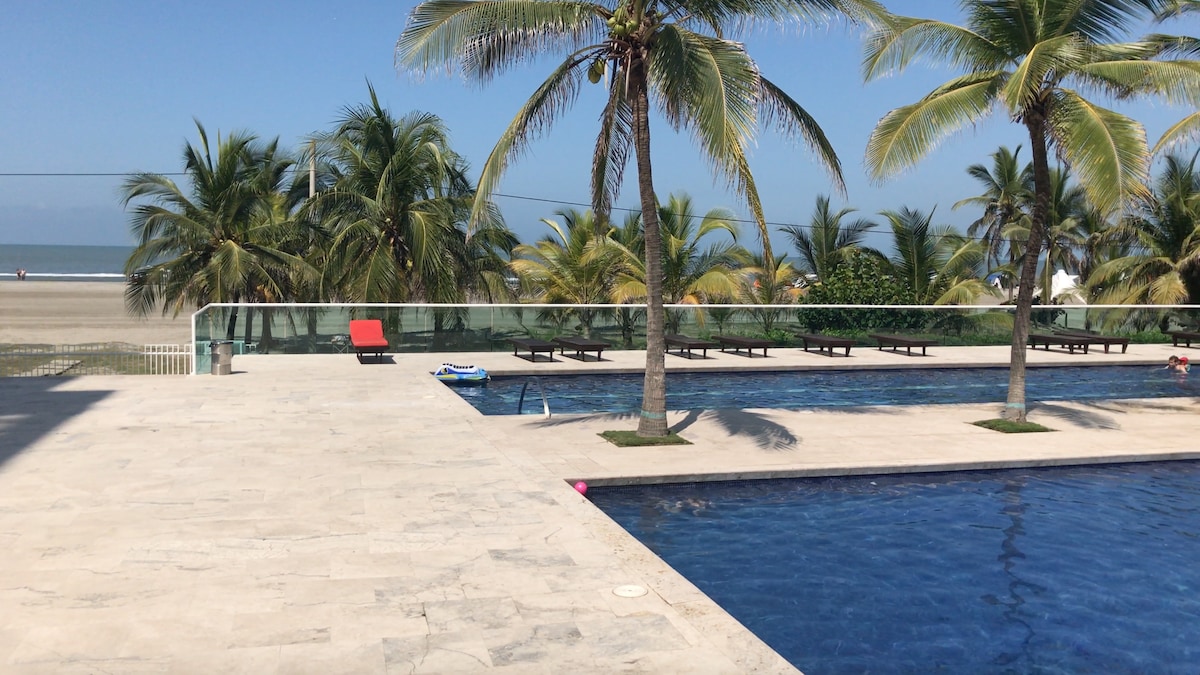 A serene outdoor pool area is visible, featuring a clear blue pool surrounded by smooth stone flooring. Palm trees provide shade, while lounge chairs line the poolside. The beach is seen in the distance, merging with the calm sea under a clear sky.