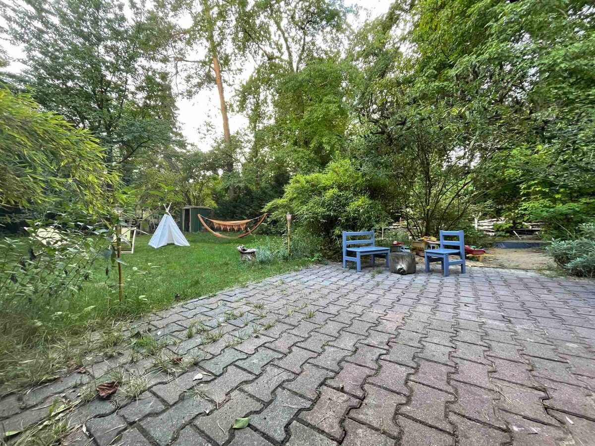 A serene garden space features a grassy area surrounded by trees, with a white tent and a hammock visible. Two blue chairs are positioned on a paved patio, offering a spot to relax and enjoy nature.