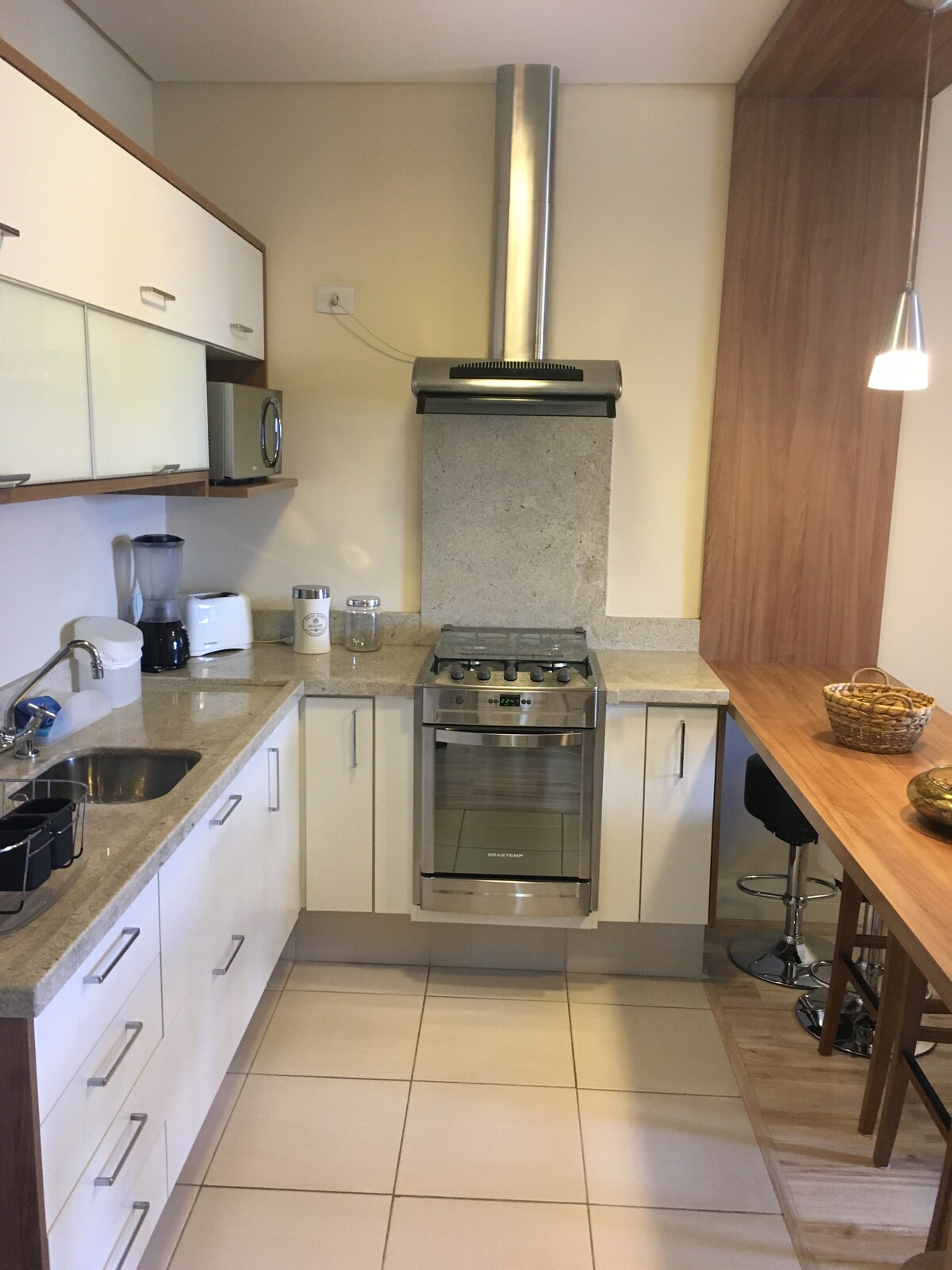 The kitchen area is equipped with modern cabinetry and a granite countertop. Stainless steel appliances include a gas stove and a microwave. A small dining table with stools provides space for meals, complemented by a fruit basket and stylish pendant lighting above.