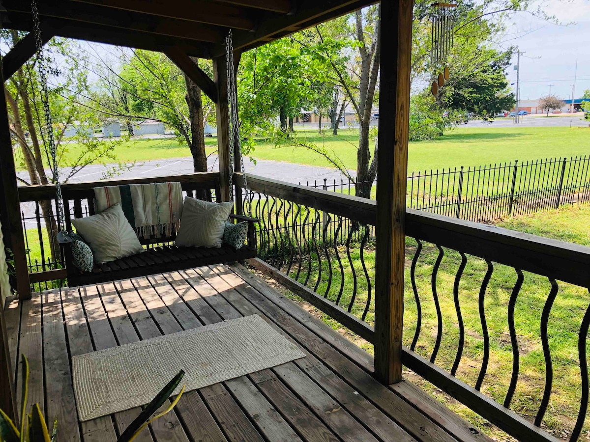 A spacious porch features a wooden swing adorned with plush cushions, complemented by a soft area rug. Sunlight filters through green foliage, casting gentle shadows on the floor. A fenced yard is visible beyond the railing, providing an open view of the surrounding greenery.