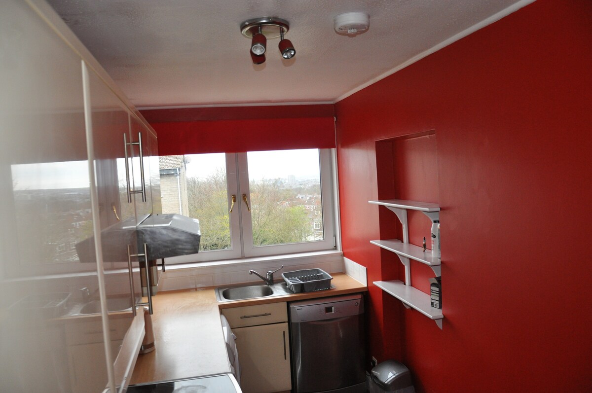 The kitchen features red walls accented by light wood countertops. A window provides natural light and a view, while cabinetry and shelves offer storage space. Appliances include a sink, dishwasher, and induction oven, with space for additional kitchenware.