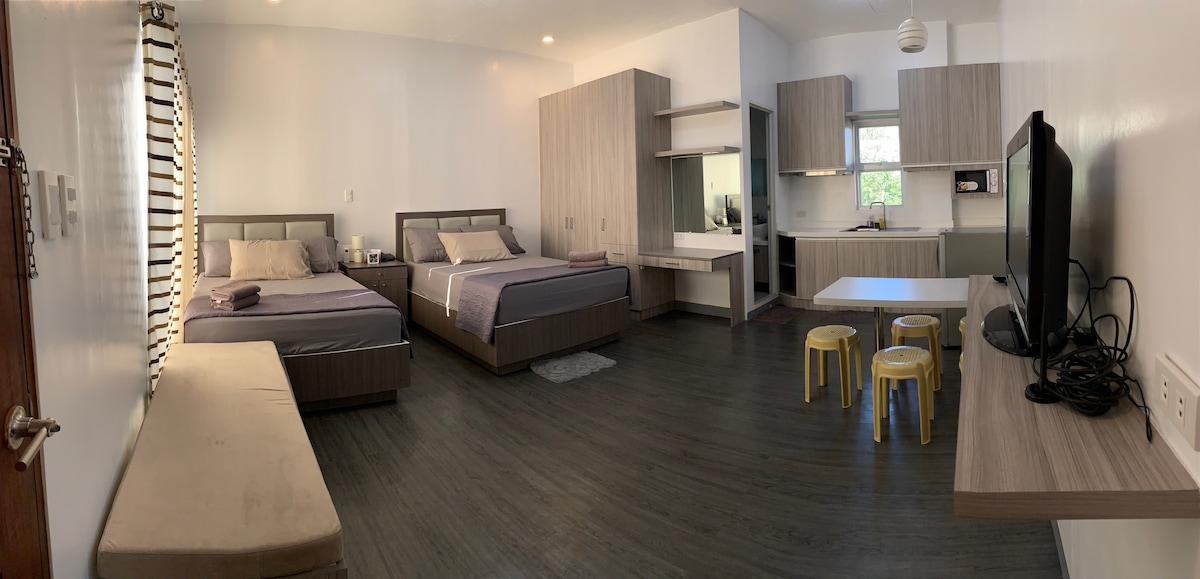 A spacious studio unit features two full double beds, a sofa bed, and a dining area with a table and chairs. A kitchenette with basic appliances is included. A 50" TV is positioned across from the seating area. Natural light enters through a window, enhancing the neutral decor.