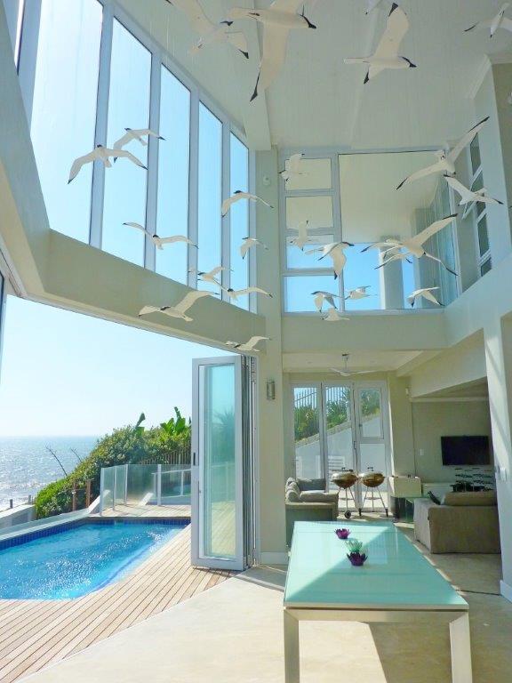 A bright and airy living area features high ceilings and large glass walls, allowing for stunning views of the ocean. A pool is visible outside, with a wooden deck surrounding it. Decorative birds hang from the ceiling, adding interest to the space.