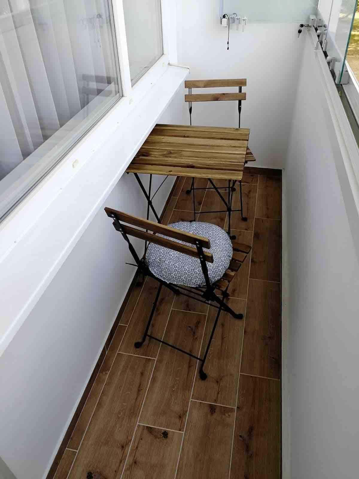 A compact balcony features a small wooden table paired with two foldable chairs. One chair is accented with a round cushion. Natural light streams in through large windows, illuminating the wooden flooring.