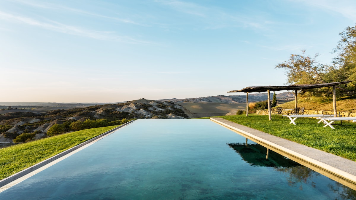 An infinity pool with a clear water surface extends towards the horizon, surrounded by lush green grass. A wooden pergola provides shaded seating, while rolling hills are visible in the background under a soft sky.
