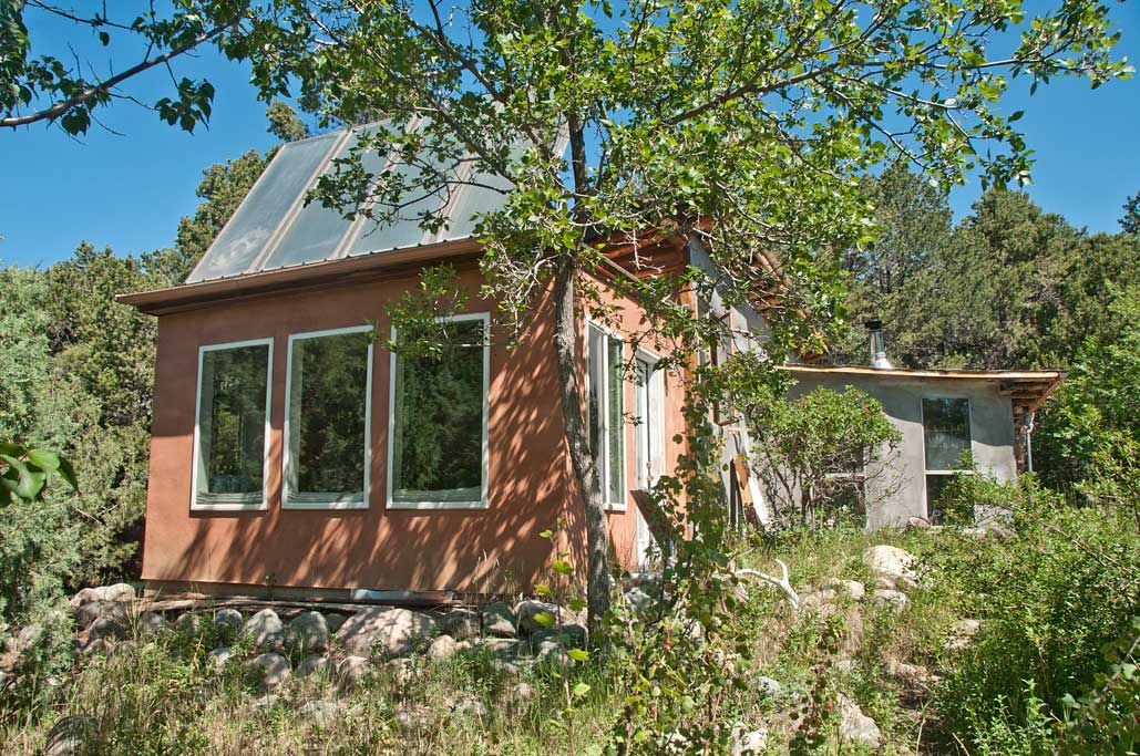 A charming rustic home is framed by trees and natural greenery. The exterior showcases a combination of earth-toned walls and large windows, allowing for ample natural light. Rocky landscaping enhances the serene environment, inviting guests to enjoy the peaceful surroundings.