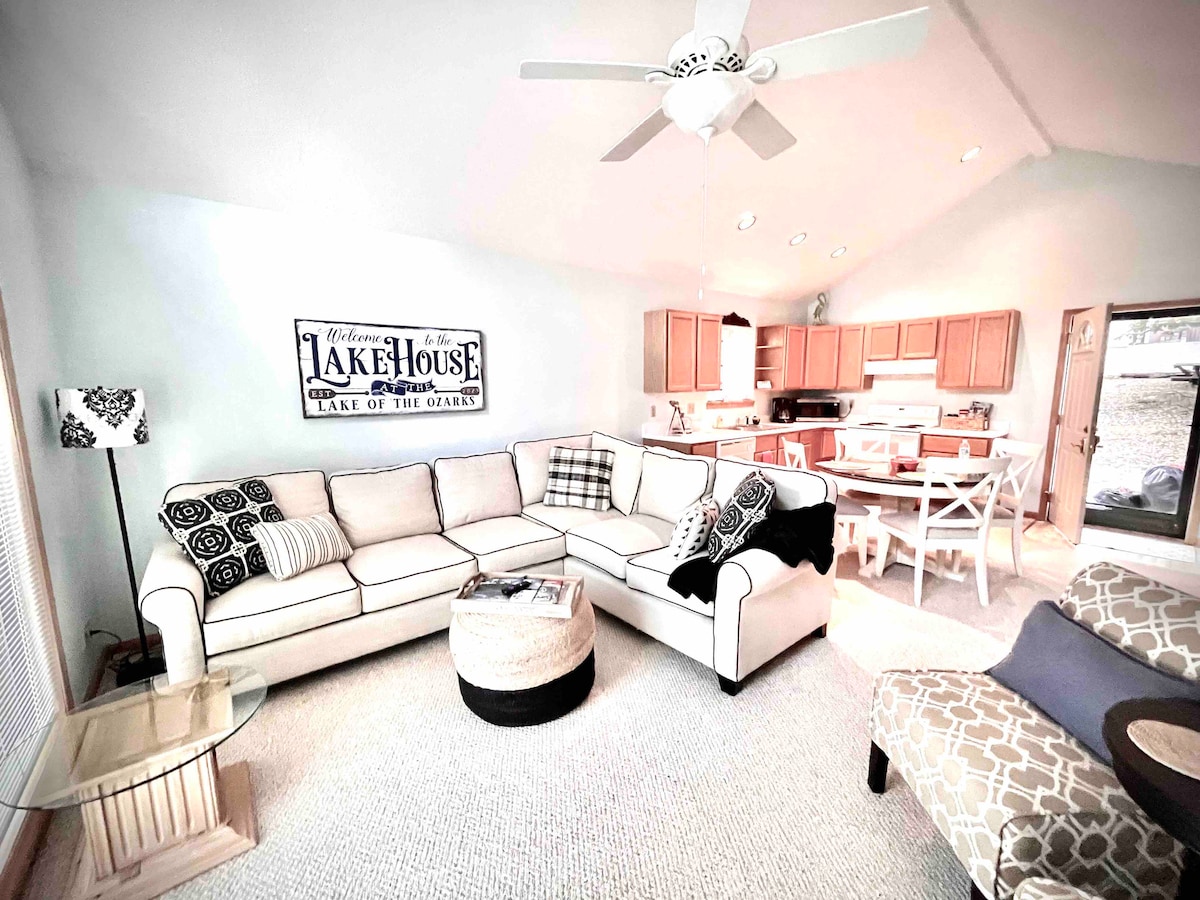 The living room features a large sectional sofa arranged around a round coffee table. Natural light fills the space through large windows, with a view of the outdoors. An open kitchen and dining area are visible in the background, highlighting a welcoming atmosphere.