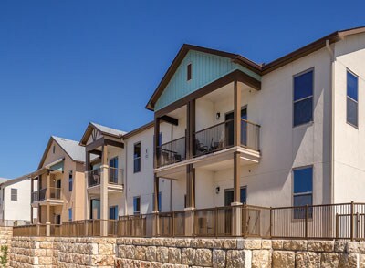 A modern two-story building is shown from an exterior perspective, featuring multiple balconies with railings. The structure is adorned with a mix of light-colored and tan siding, complemented by blue accents and large windows reflecting sunlight under a clear blue sky.