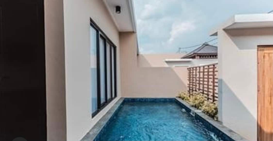 A private pool is surrounded by a stone deck, featuring clear blue water under natural light. Large windows frame the pool area, allowing for a pleasing view while providing a sense of privacy. Lush greenery is visible along the perimeter.
