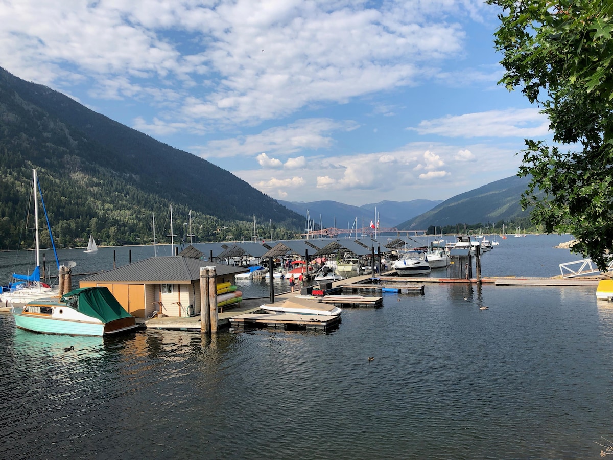 A serene waterfront view features a marina filled with various boats and sailboats, nestled between lush green mountains. The sky is partially cloudy, reflecting soft light on the water's surface, creating a tranquil atmosphere for relaxation and enjoyment.