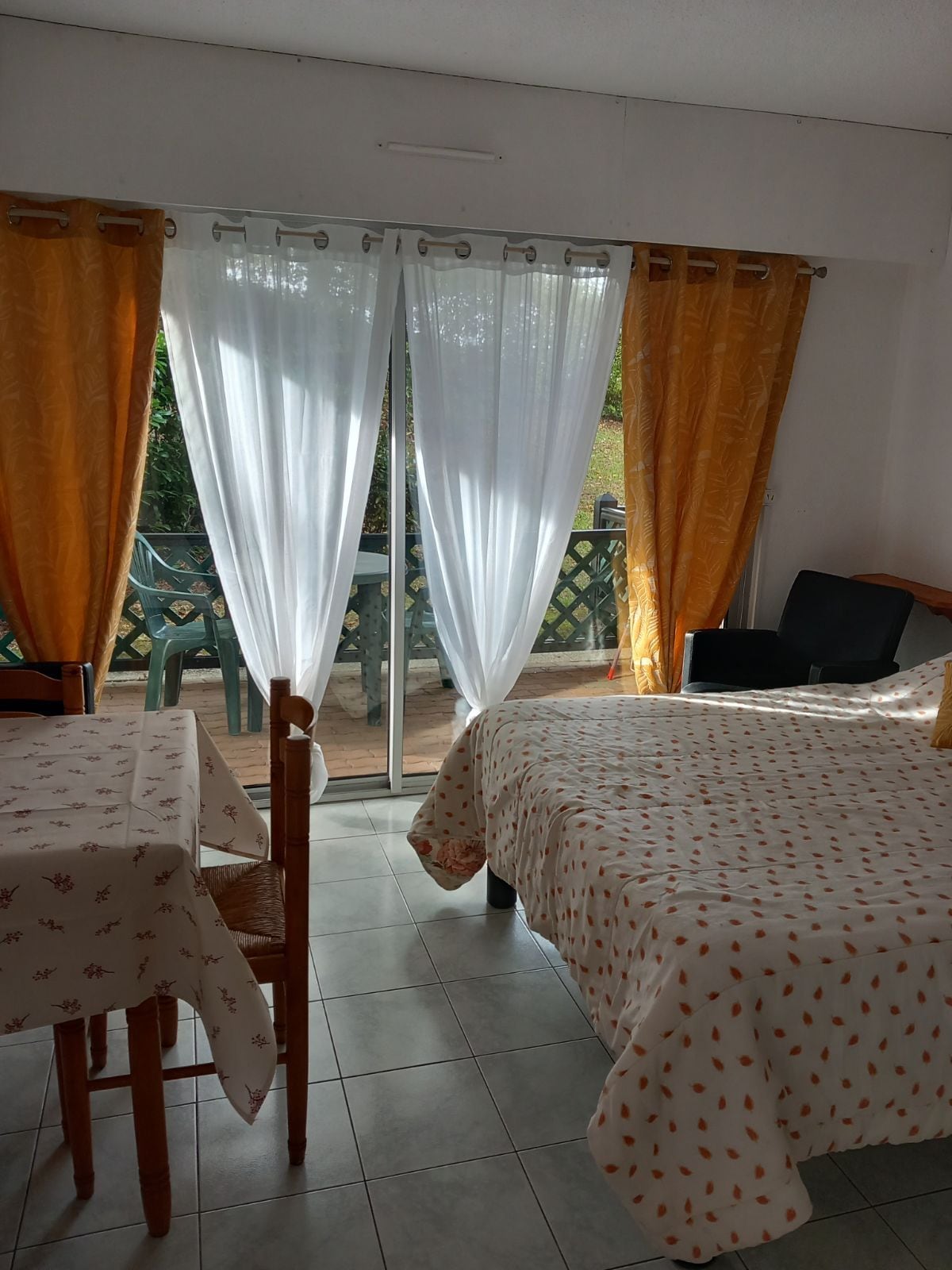 A bright interior features large windows with sheer white and golden drapes, allowing natural light to filter through. A spacious bed is covered with a patterned bedspread, accompanied by a small dining table and chairs. A patio area is visible through the glass doors.