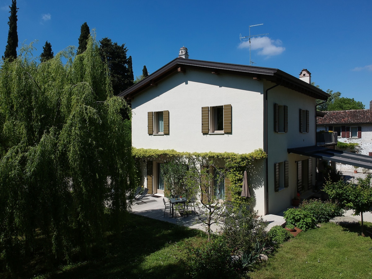 A two-story house is surrounded by a lush garden, featuring green climbing plants along the exterior walls. Windows with wooden shutters allow natural light to fill the interior. A patio area includes a table and chairs, inviting outdoor relaxation amidst the serene landscape.