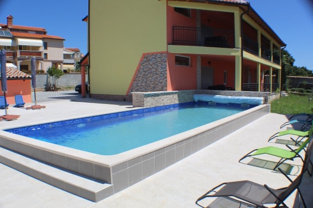 The outdoor area features a spacious saltwater pool with a waterfall and massage jets, surrounded by relaxing lounge chairs. The building consists of bright colored walls, with an accessible entrance to upper-level apartments visible in the background. Clear skies contribute to a sunny ambiance.