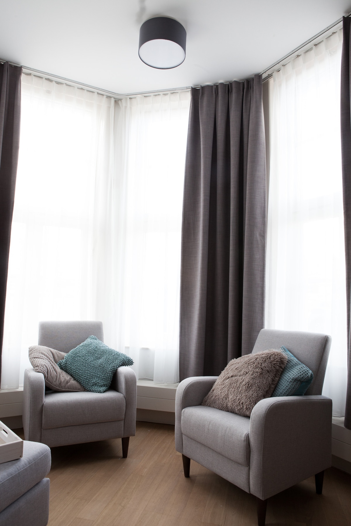 A bright seating area is defined by large windows draped with sheer curtains, allowing natural light to fill the space. Two inviting armchairs are positioned in the corner, each adorned with textured cushions for added comfort. Neutral tones of the furniture create a serene ambiance.