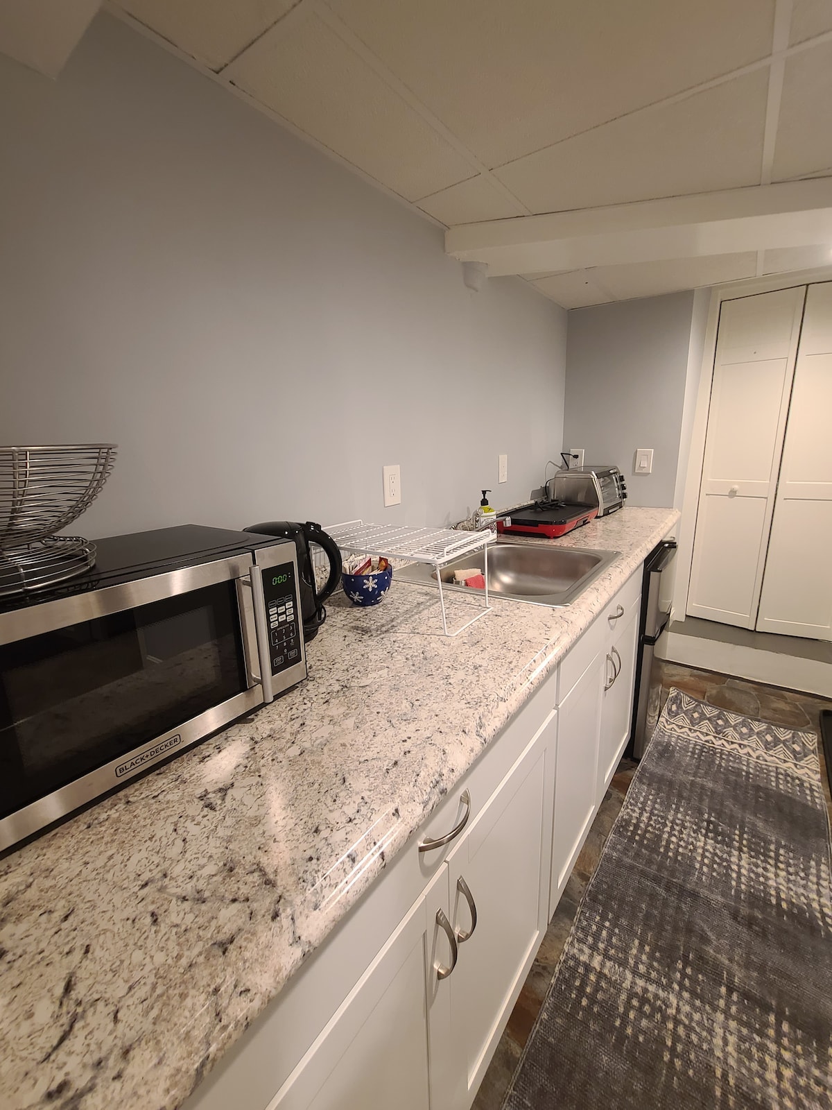 The kitchenette features modern appliances including a microwave, kettle, and small fridge. Stone countertops offer ample preparation space. Cabinets provide storage, and a small dish rack is placed on the counter, enhancing functionality in this cozy area.