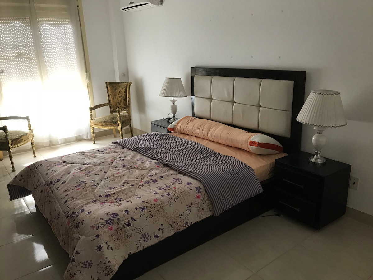 A spacious bedroom features a large bed with a light-colored blanket and decorative pillows. Two bedside tables with lamps are positioned on either side of the bed. A pair of elegant chairs sit beside a window, allowing natural light to enter the room.