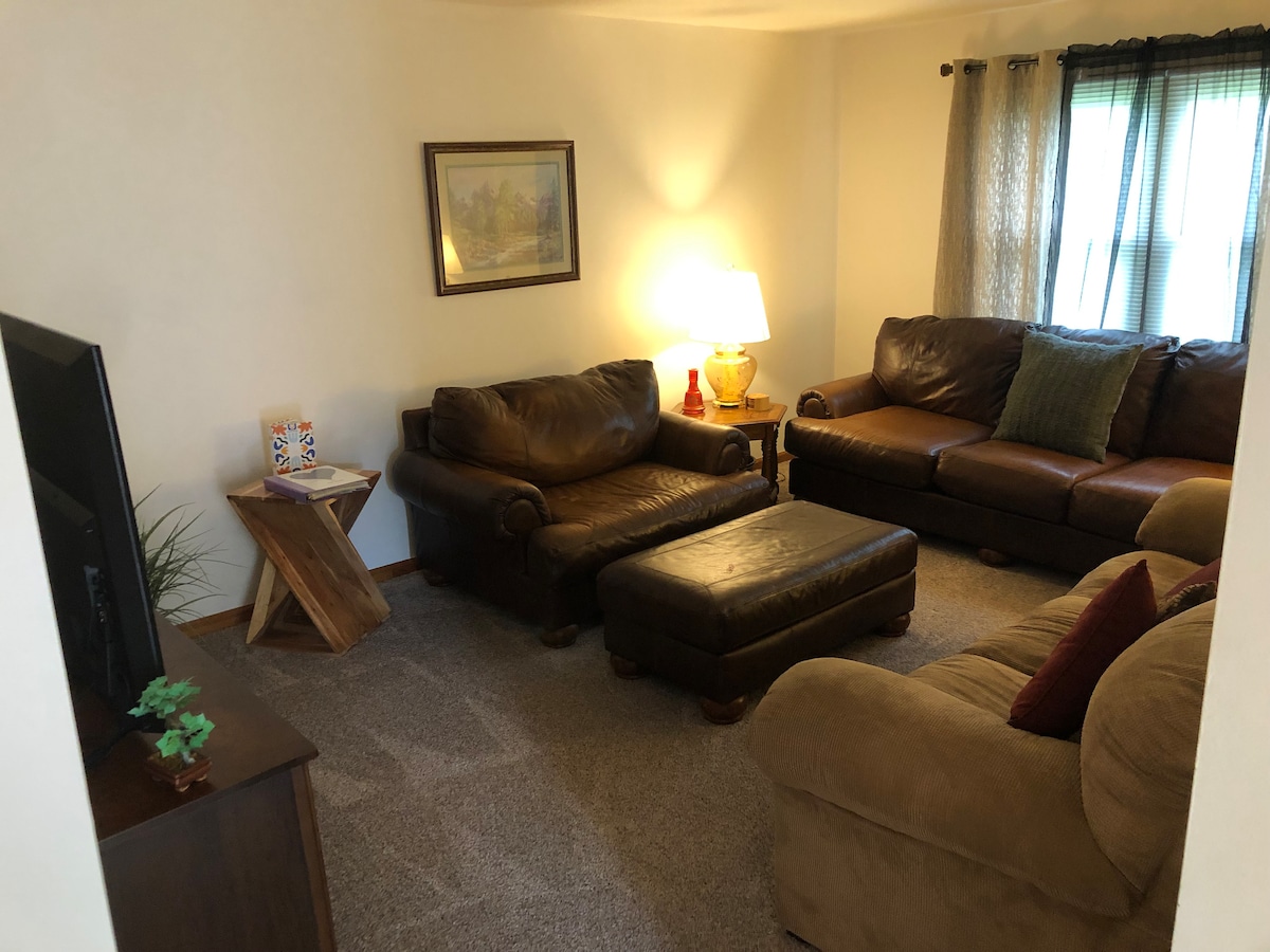 A comfortable living area is furnished with two brown leather couches and a tan couch topped with decorative pillows. A wooden coffee table sits at the center. Natural light enters through the window, complemented by soft curtains. A television is positioned on a dark wooden stand.